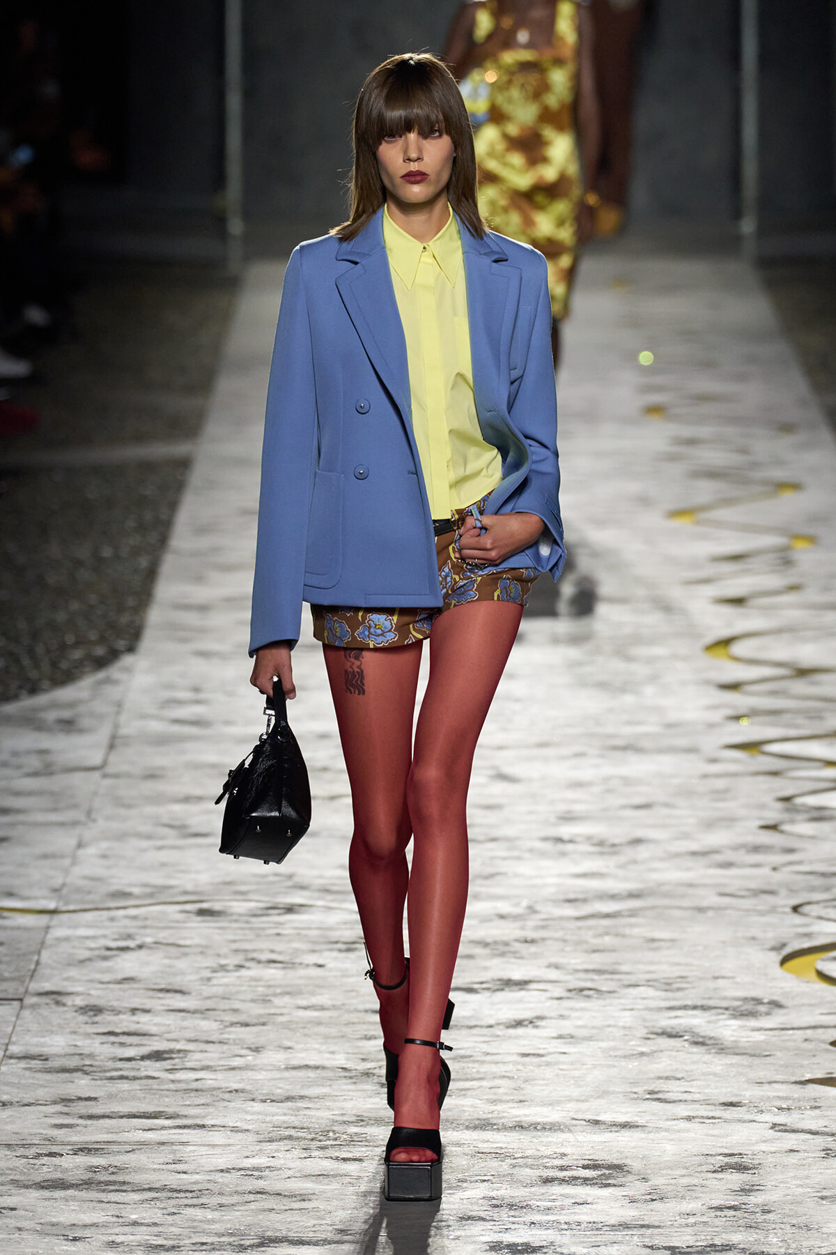 Model walking the runway in a light blue oversized blazer over a pale yellow shirt, paired with floral shorts, red tights, black platform sandals, and a small black handbag.