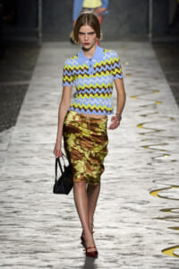 Model walking a fashion runway in a zigzag print short-sleeve polo and a satin floral midi skirt, carrying a black handbag.