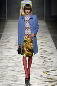 Model struts a runway in a light blue coat over a lavender zigzag top and metallic olive-green floral skirt, with black platform sandals and a small black bag.