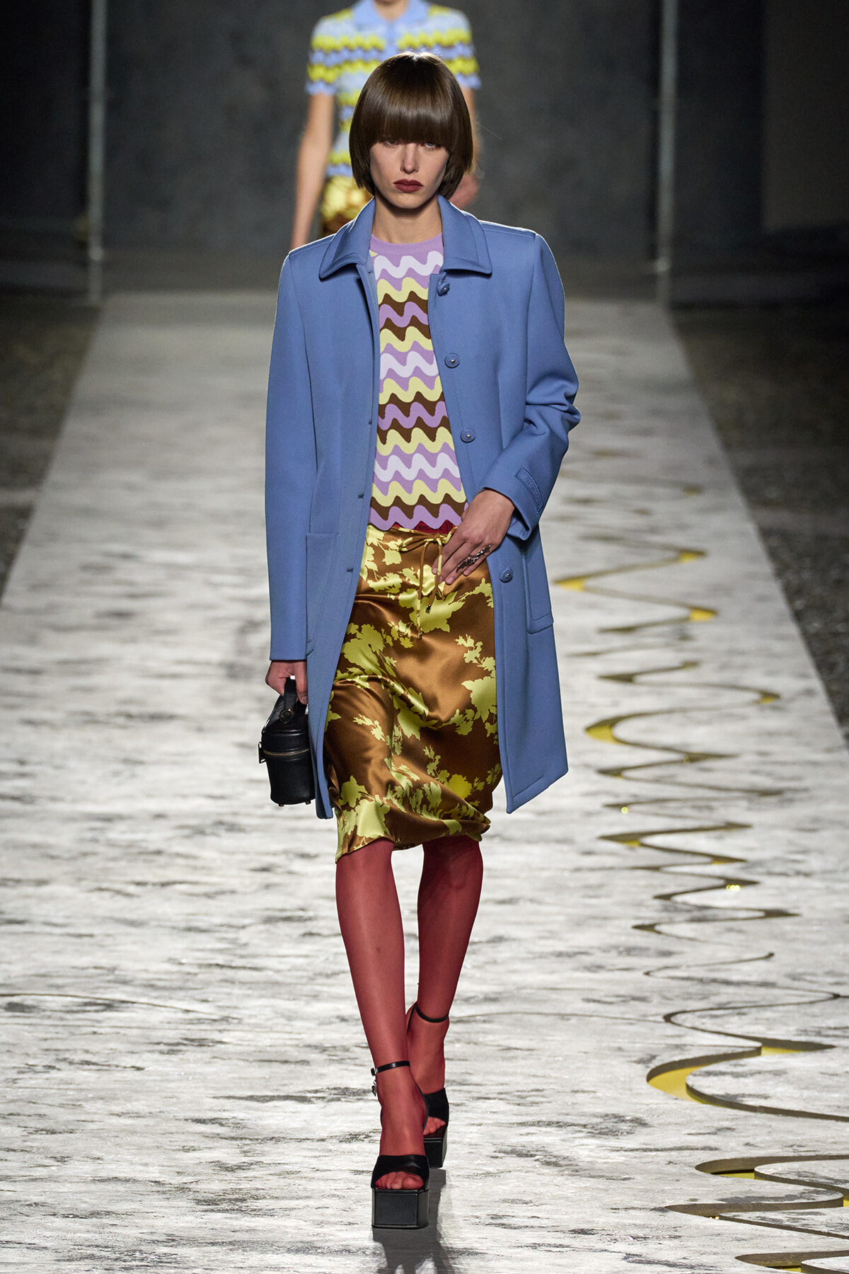 Model struts a runway in a light blue coat over a lavender zigzag top and metallic olive-green floral skirt, with black platform sandals and a small black bag.