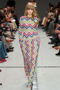 Model walking the runway in a full-length multicolor zigzag-print jumpsuit with a geometric accessory at the shoulder, audience on both sides.