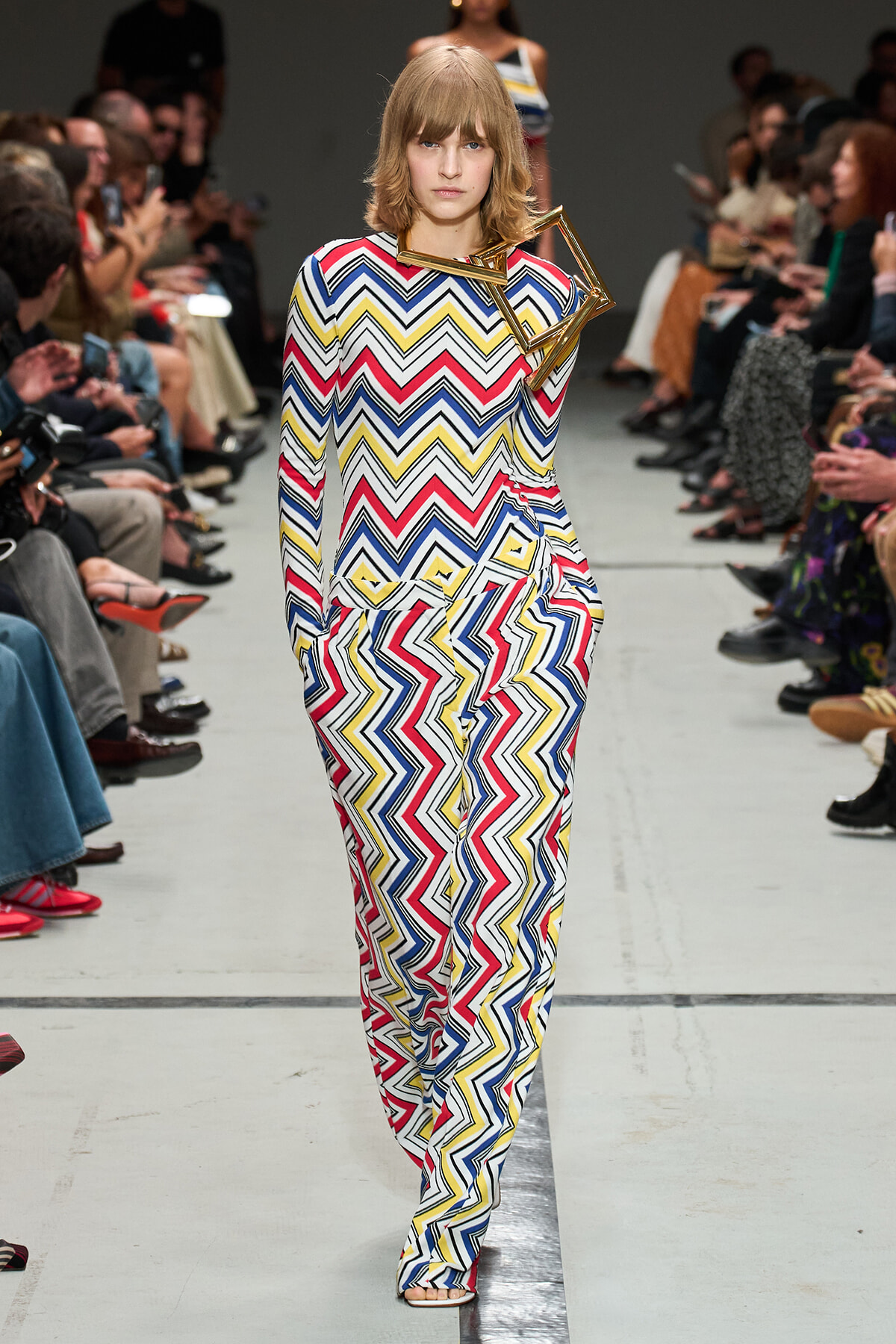 Model walking the runway in a full-length multicolor zigzag-print jumpsuit with a geometric accessory at the shoulder, audience on both sides.