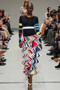 Runway model wearing a black form-fitting dress with color-band sleeves and a bold multicolor zigzag ruffled skirt, paired with gold neck rings and yellow sandals.