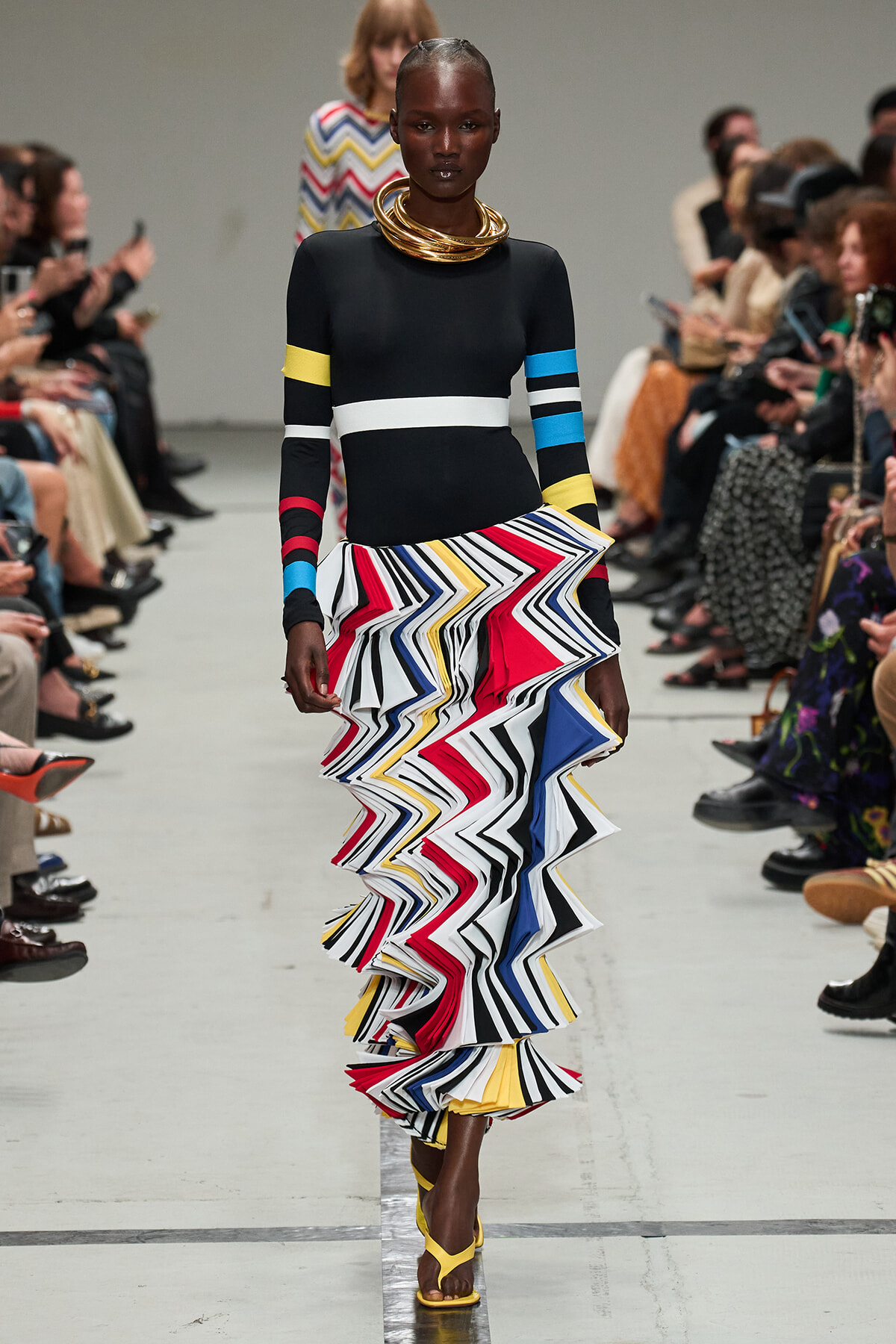 Runway model wearing a black form-fitting dress with color-band sleeves and a bold multicolor zigzag ruffled skirt, paired with gold neck rings and yellow sandals.