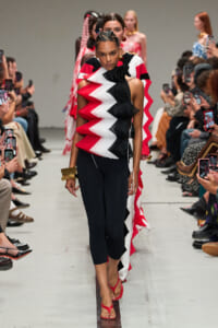 Model walks a fashion show in a red, white, and black zigzag ruffled top with black leggings and red sandals on a runway audience filming nearby.