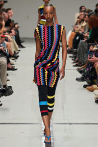 Model walking a runway in a sleeveless dress with a bright zigzag pattern, paired with black leggings featuring bold color bands and blue sandals.