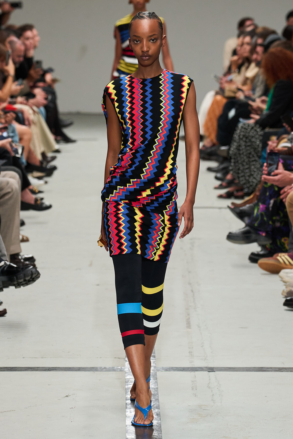 Model walking a runway in a sleeveless dress with a bright zigzag pattern, paired with black leggings featuring bold color bands and blue sandals.