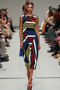Model walking a runway in a sleeveless, multicolor striped dress with bold diagonal panels and a chunky gold necklace, red sandals visible at the bottom.