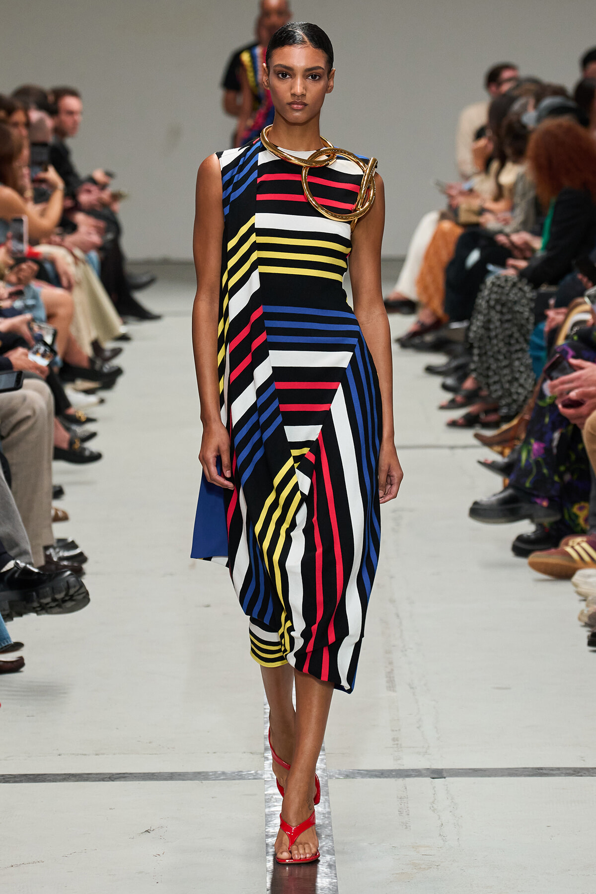 Model walking a runway in a sleeveless, multicolor striped dress with bold diagonal panels and a chunky gold necklace, red sandals visible at the bottom.