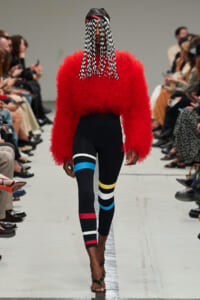 Model walks a runway in a bright red fuzzy sweater and black leggings with multicolored bands; hair features long black-and-white beaded strands over the face.