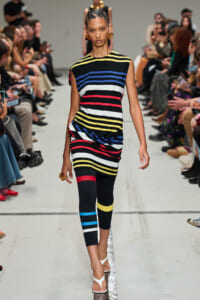 Model on a runway wearing a color-blocked striped dress and matching leggings; audience seated along the sides.