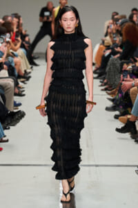 Model walks a runway in a black layered ruffle sleeveless gown with sheer panels, audience on both sides.Jewelry includes bold gold earrings and rectangular bracelets on her wrists.