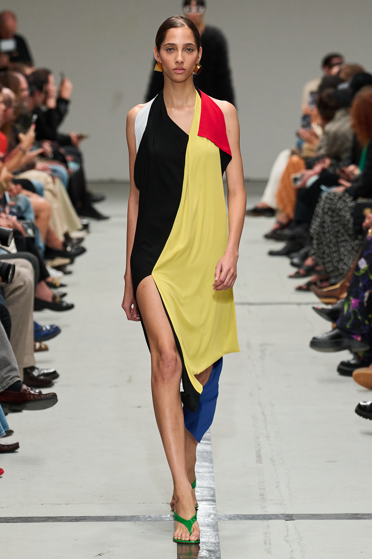 Model walks a fashion runway in a color-block sleeveless dress with black, yellow, red, and white panels and a thigh-high slit; green sandals.