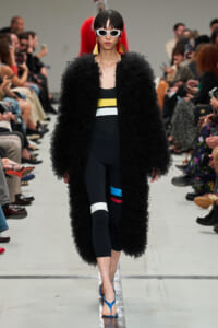 Model walking a runway in a long black fluffy coat over a color-blocked black outfit; white sunglasses and bold yellow earrings, blue sandals; audience watches.
