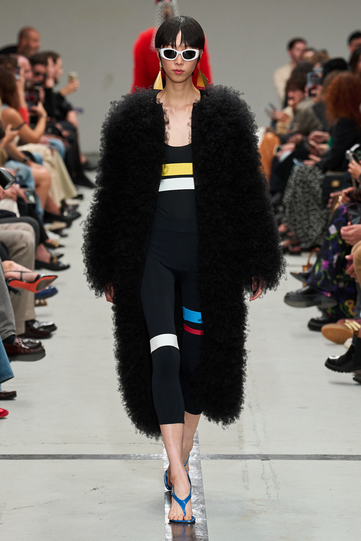 Model walking a runway in a long black fluffy coat over a color-blocked black outfit; white sunglasses and bold yellow earrings, blue sandals; audience watches.
