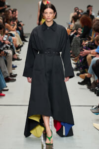 Model walking runway in a long black belted coat-dress with oversized sleeves and a multicolored inner hem; green sandals visible.