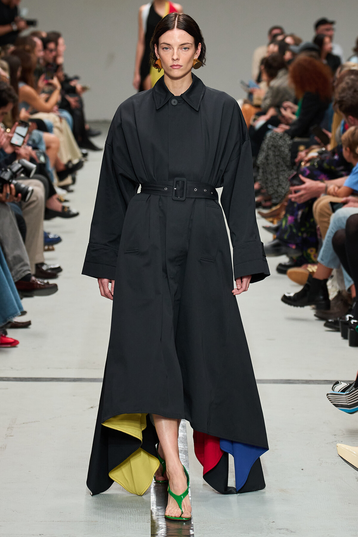 Model walking runway in a long black belted coat-dress with oversized sleeves and a multicolored inner hem; green sandals visible.