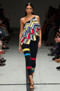 Model on a runway wearing a one-shoulder zigzag ruffle top in black, white, yellow, blue, red with color-block leggings and sandals.
