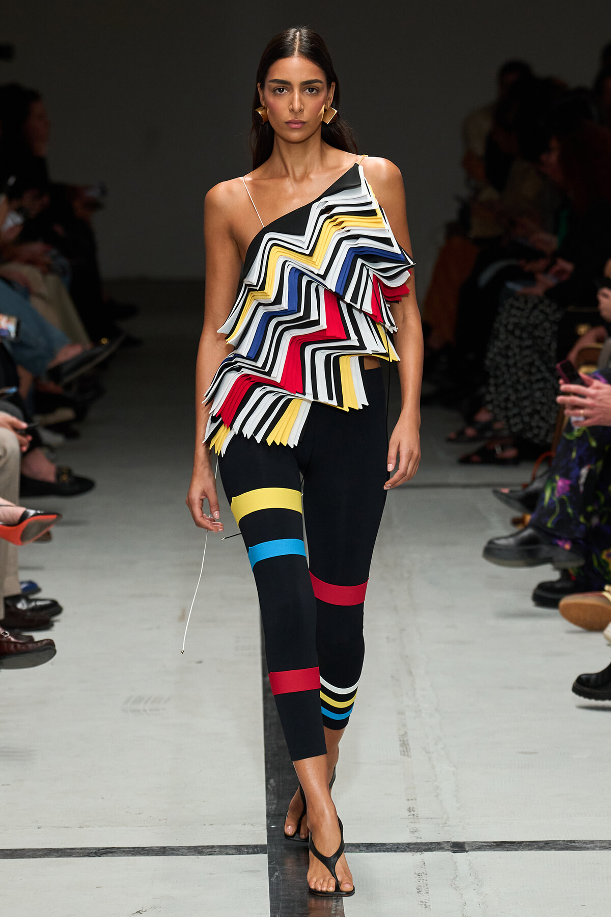 Model on a runway wearing a one-shoulder zigzag ruffle top in black, white, yellow, blue, red with color-block leggings and sandals.