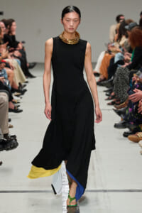 Model on a runway wearing a sleeveless black dress with a color-blocked, layered hem and green sandals; gold stacked necklace accents the look.