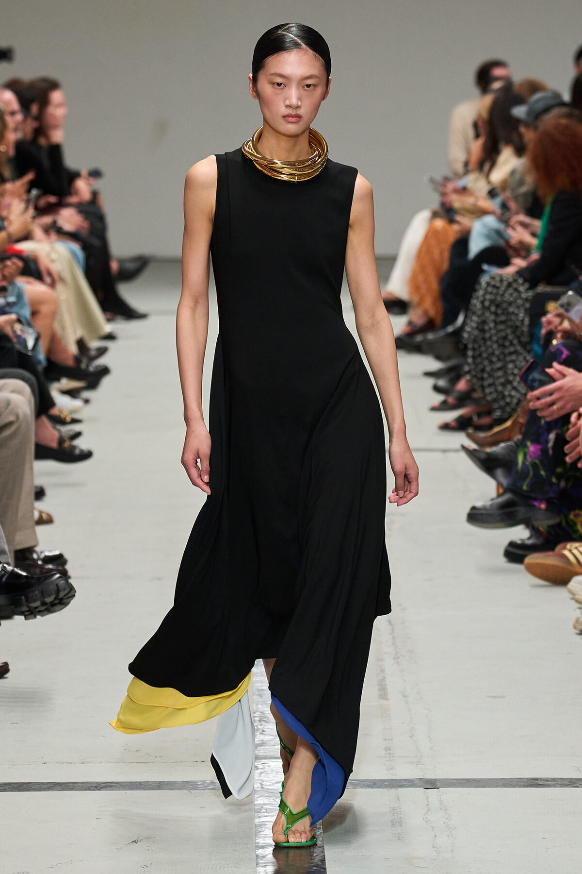Model on a runway wearing a sleeveless black dress with a color-blocked, layered hem and green sandals; gold stacked necklace accents the look.