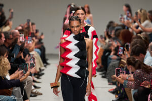 Model walks the runway wearing a bold red‑white‑black zigzag top; audience lines up filming with smartphones on both sides.