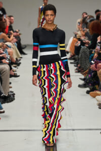 Model on a fashion show runway wearing a black top with color bands and a bright, multi-color zigzag ruffle skirt.