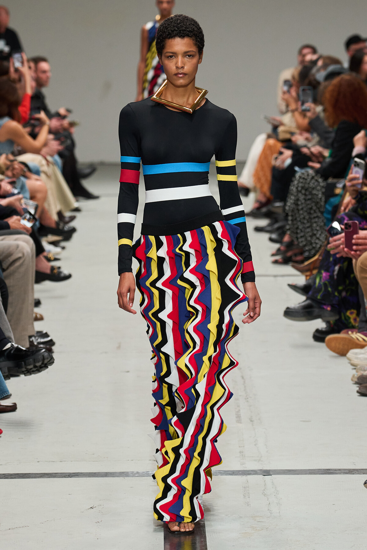 Model on a fashion show runway wearing a black top with color bands and a bright, multi-color zigzag ruffle skirt.