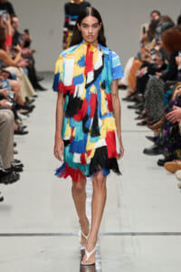 Model walks the runway in a vibrant, multicolor fringe dress with a blue top and white sandals at a fashion show