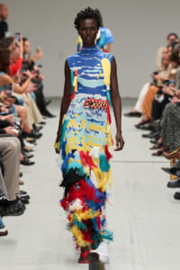 Model walking runway in sleeveless blue dress with colorful abstract print and long multicolored fringe tassels.