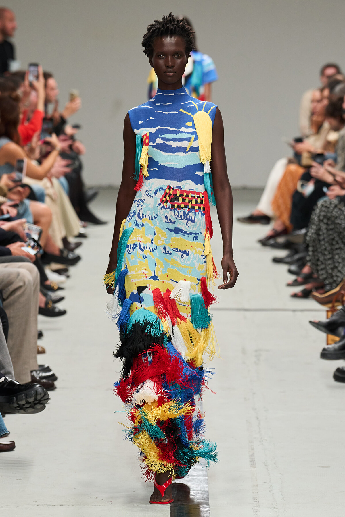 Model walking runway in sleeveless blue dress with colorful abstract print and long multicolored fringe tassels.