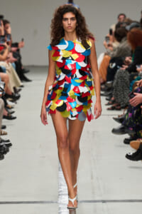 Model walks a runway in a short dress made of overlapping colorful circular patches of red, blue, yellow, white, and black, with white sandals.