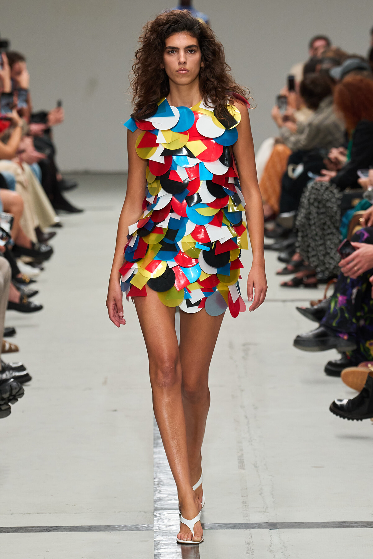 Model walks a runway in a short dress made of overlapping colorful circular patches of red, blue, yellow, white, and black, with white sandals.