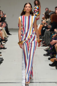 Model on runway wearing a white zigzag-pattern jumpsuit in red, blue, yellow with gold geometric earrings and cuffs in view of the audience.