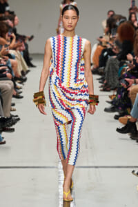 Model walks the runway wearing a sleeveless, zigzag-pattern jumpsuit in red, blue, yellow and white with matching leg-length pants; gold cuffs and yellow sandals add metallic accents.