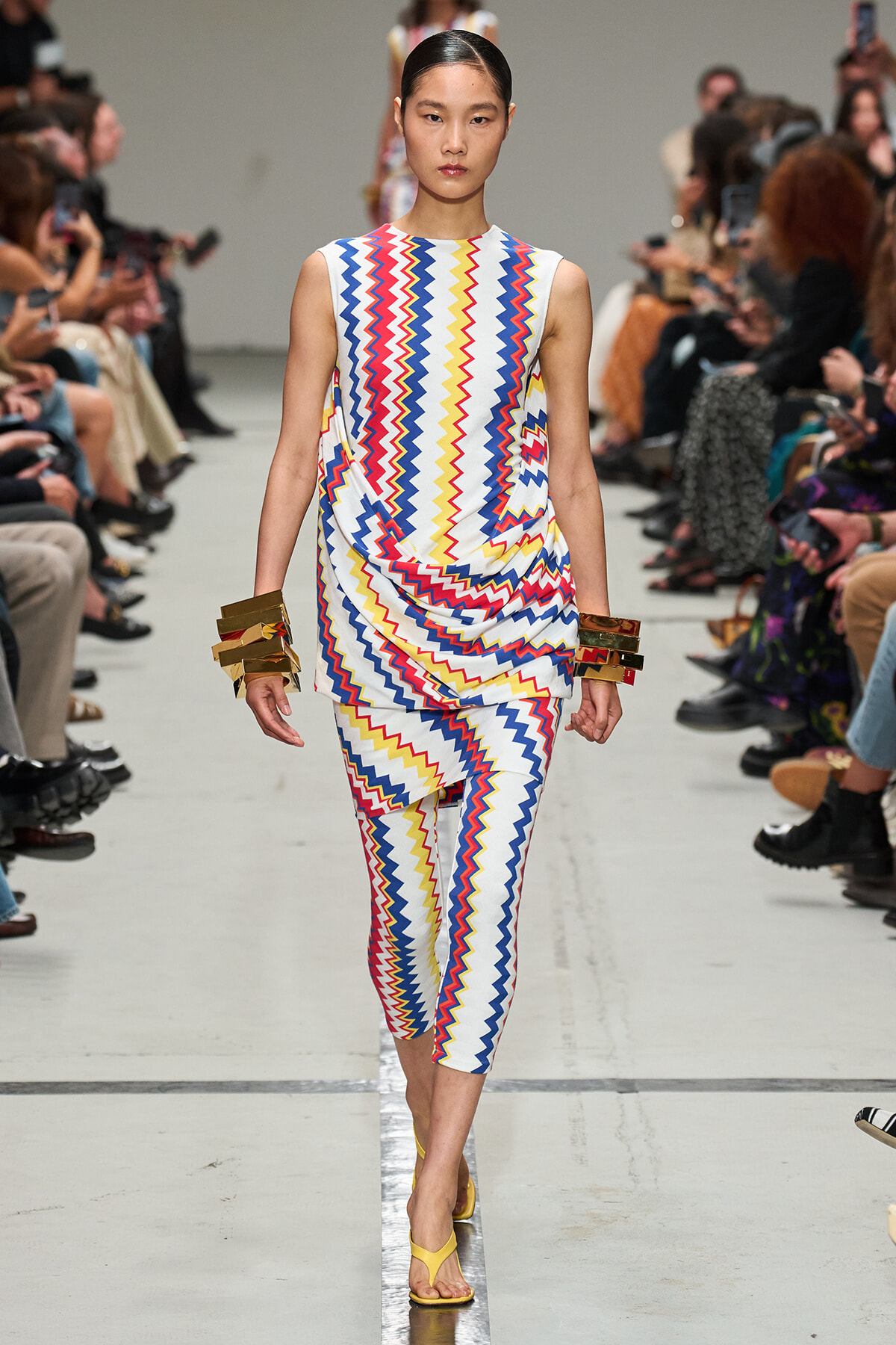 Model walks the runway wearing a sleeveless, zigzag-pattern jumpsuit in red, blue, yellow and white with matching leg-length pants; gold cuffs and yellow sandals add metallic accents.