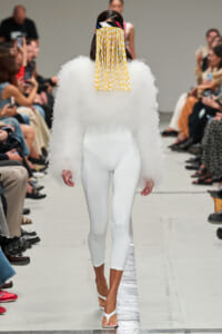 Model walks runway in all-white ensemble: fluffy textured top, tight white leggings, white sandals, and a colorful fringe headpiece.
