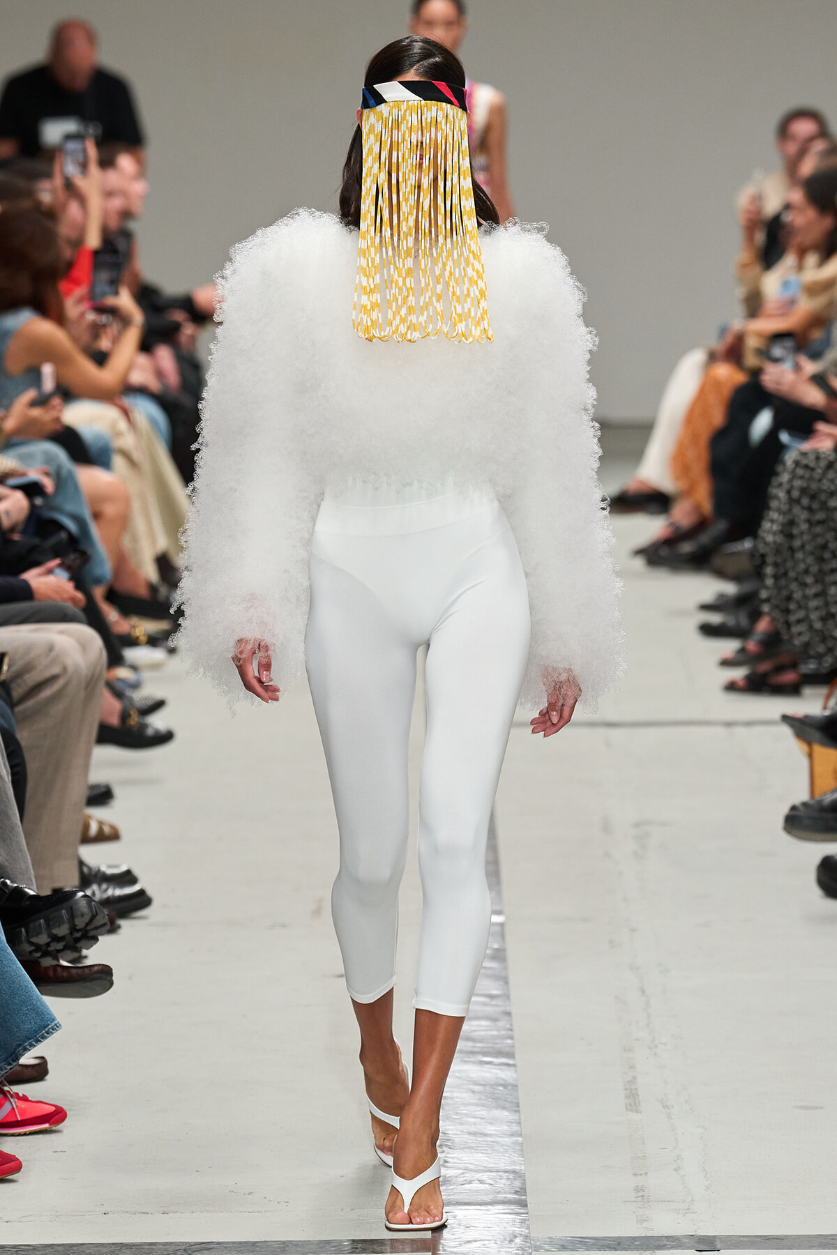 Model walks runway in all-white ensemble: fluffy textured top, tight white leggings, white sandals, and a colorful fringe headpiece.