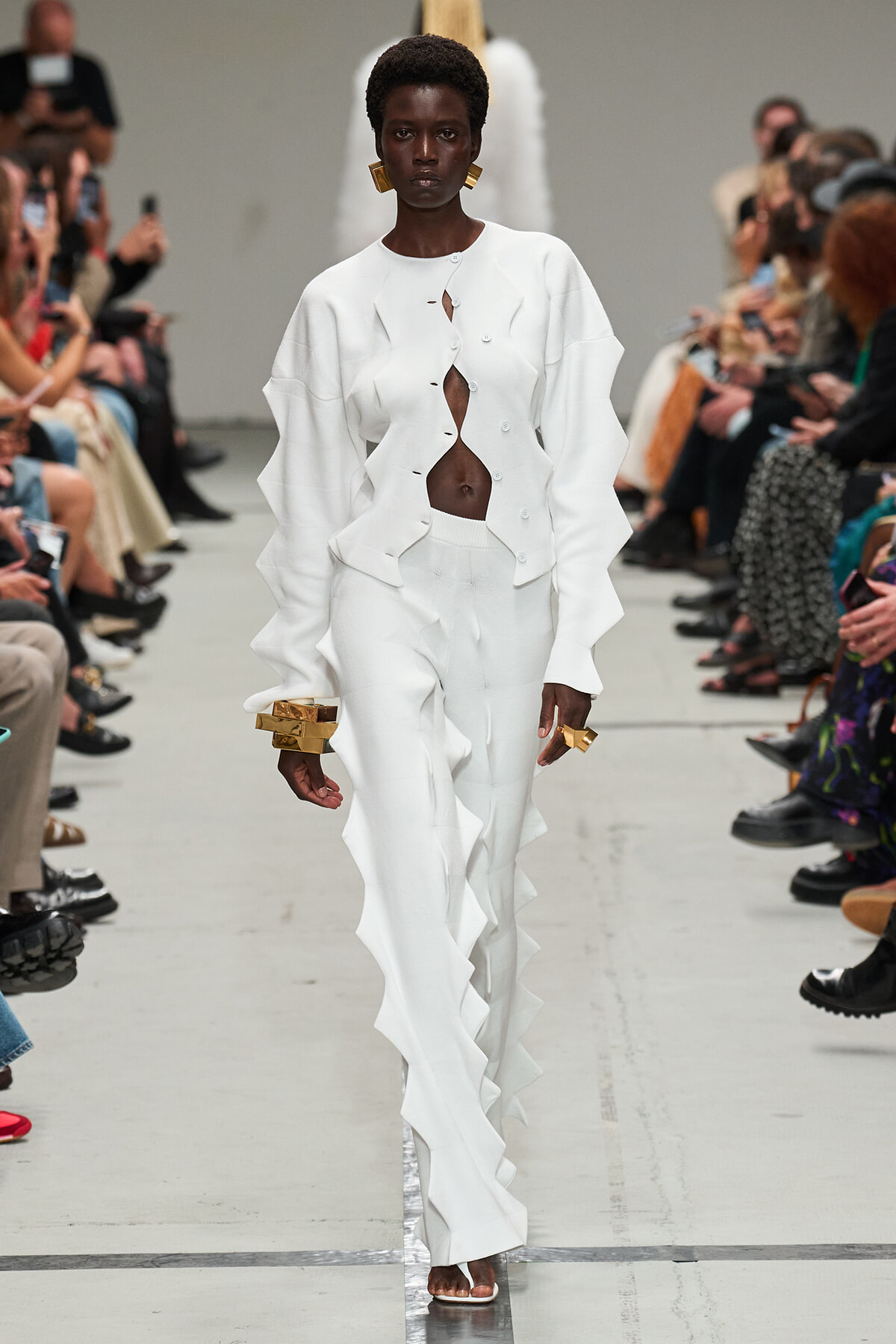 Runway model in an all-white outfit with a cut-out cardigan and ruffled panels, walking the show with large gold bracelets.