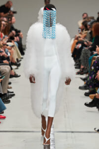 Model walks a runway in an all-white outfit: long fluffy coat over white bodysuit, blue fringe headpiece, and white heeled sandals, with a seated audience on both sides.