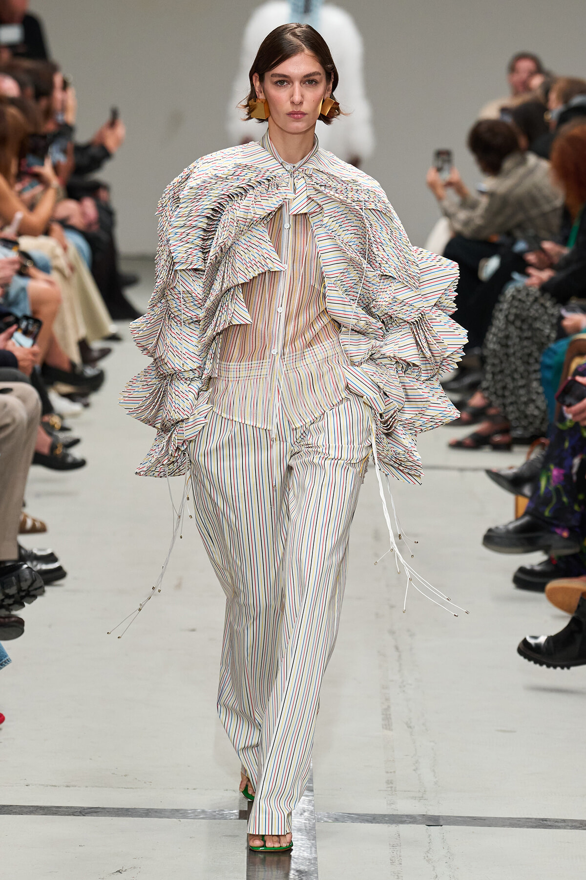 Model walks the runway in a matching striped suit with dramatic layered ruffles cascading over shoulders and arms, during a fashion show.