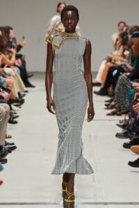 Model on a runway wearing a sleeveless black-and-white zigzag (chevron) floor-length dress with a gold geometric shoulder/neck accessory, surrounded by seated spectators.