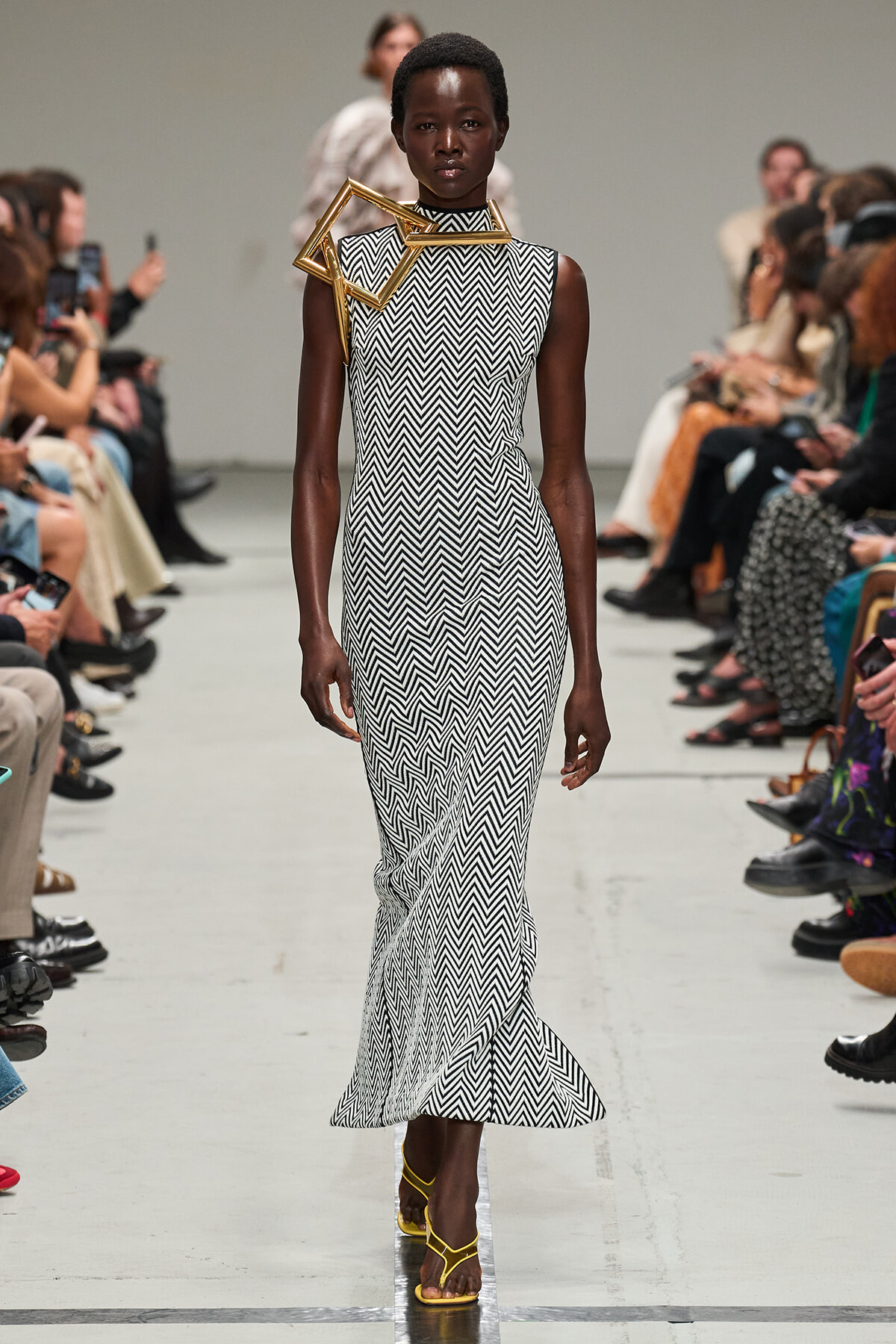 Model on a runway wearing a sleeveless black-and-white zigzag (chevron) floor-length dress with a gold geometric shoulder/neck accessory, surrounded by seated spectators.