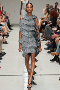 Models a black-and-white zigzag ruffle dress walking the runway; asymmetrical shoulders, high-low hem, onlookers in the background.