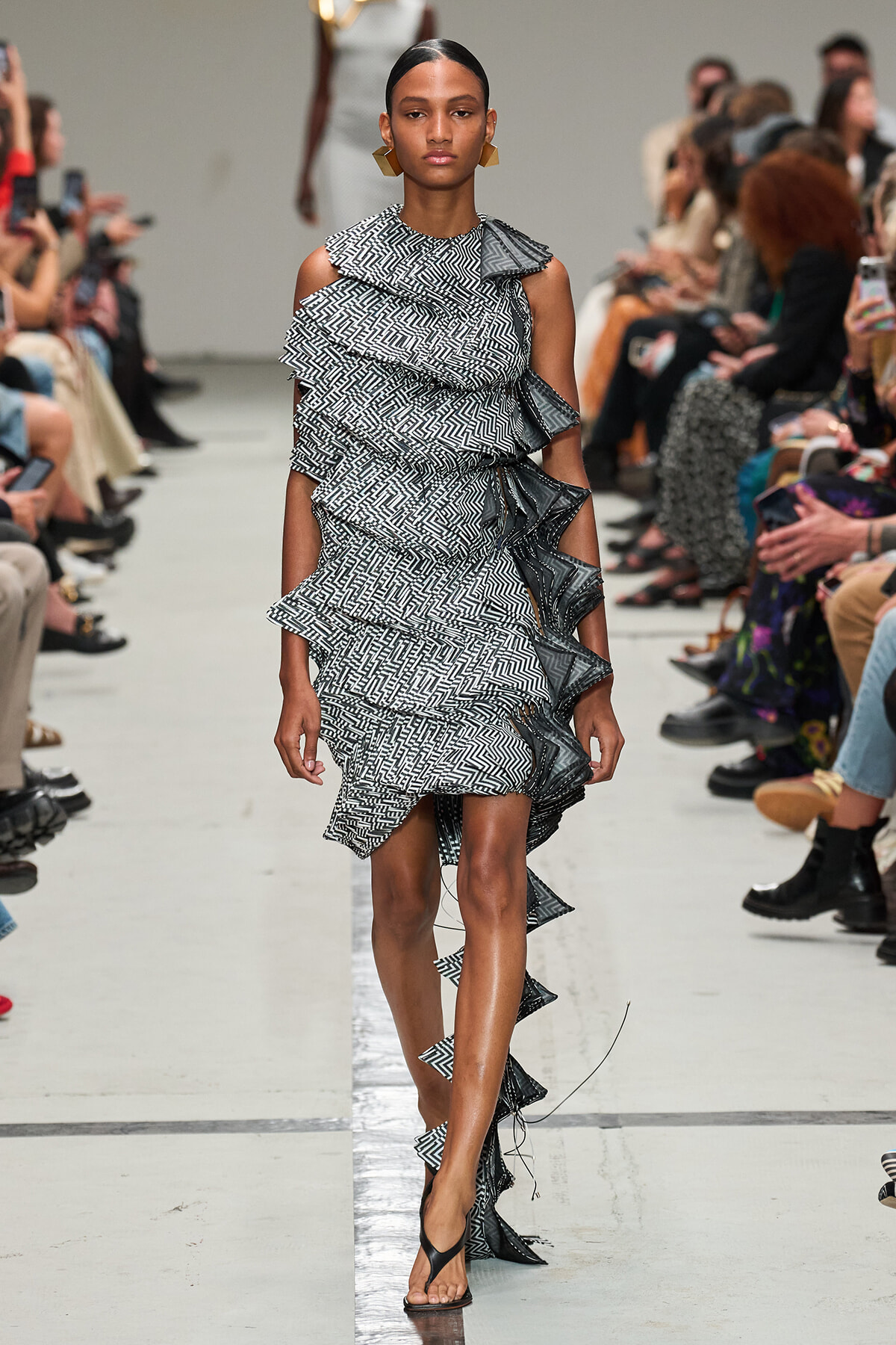 Models a black-and-white zigzag ruffle dress walking the runway; asymmetrical shoulders, high-low hem, onlookers in the background.