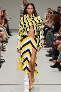 Model walking a runway in a yellow, black and white zigzag dress with a dramatic ruffled top and high-slit skirt, yellow sandals.