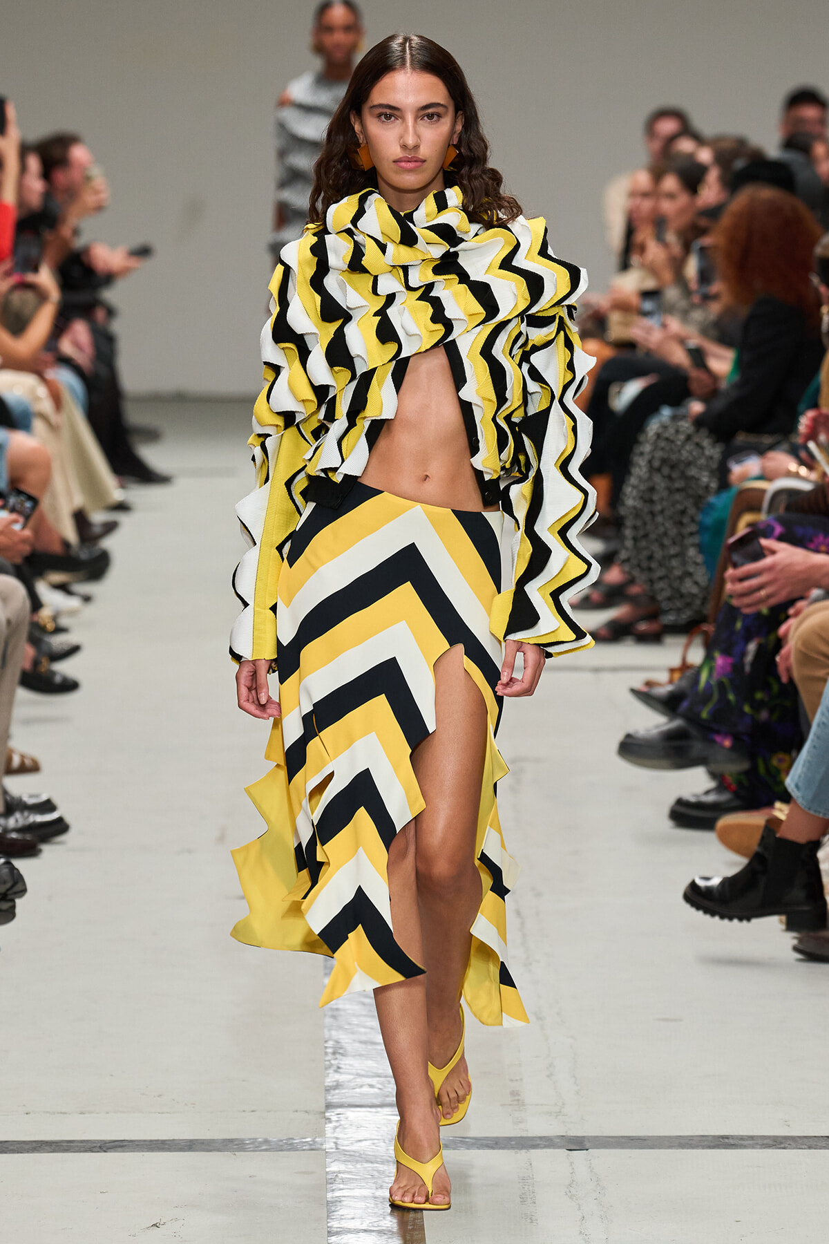 Model walking a runway in a yellow, black and white zigzag dress with a dramatic ruffled top and high-slit skirt, yellow sandals.