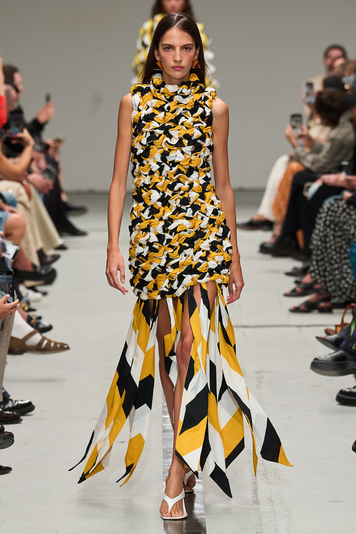 Runway model in a sleeveless yellow, black, and white textured dress with knot-like braiding and long geometric fringe at the hem, white sandals.