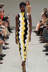 Model walking a runway in a sleeveless black, white, and yellow zigzag dress with yellow sandals and a blue headband.