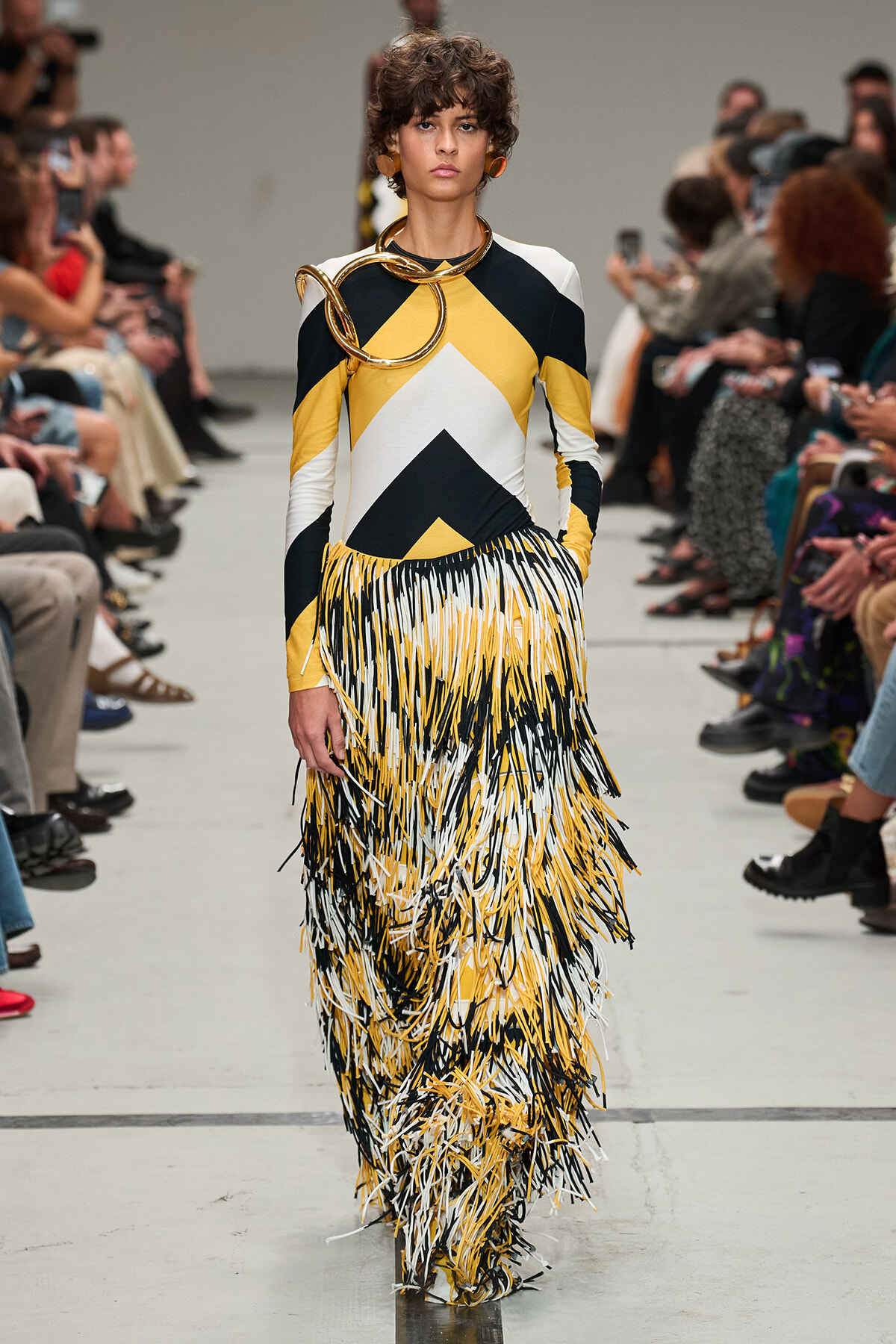 Model walks the runway in a color-block dress (yellow, black, white) with a long fringe skirt and oversized gold neck accessories.